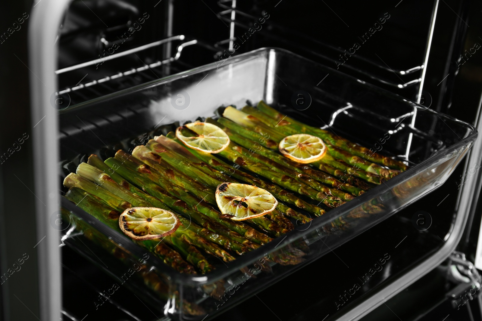 Photo of Cooked asparagus with lemon slices in glass baking dish on oven rack, closeup