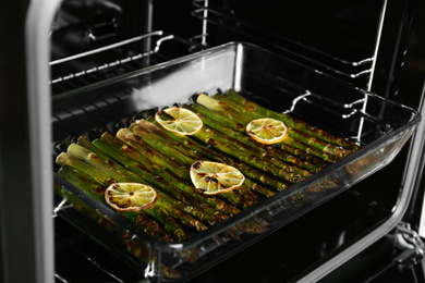 Photo of Cooked asparagus with lemon slices in glass baking dish on oven rack, closeup