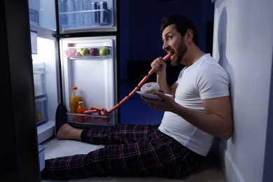 Photo of Man eating sausages near refrigerator in kitchen at night. Bad habit