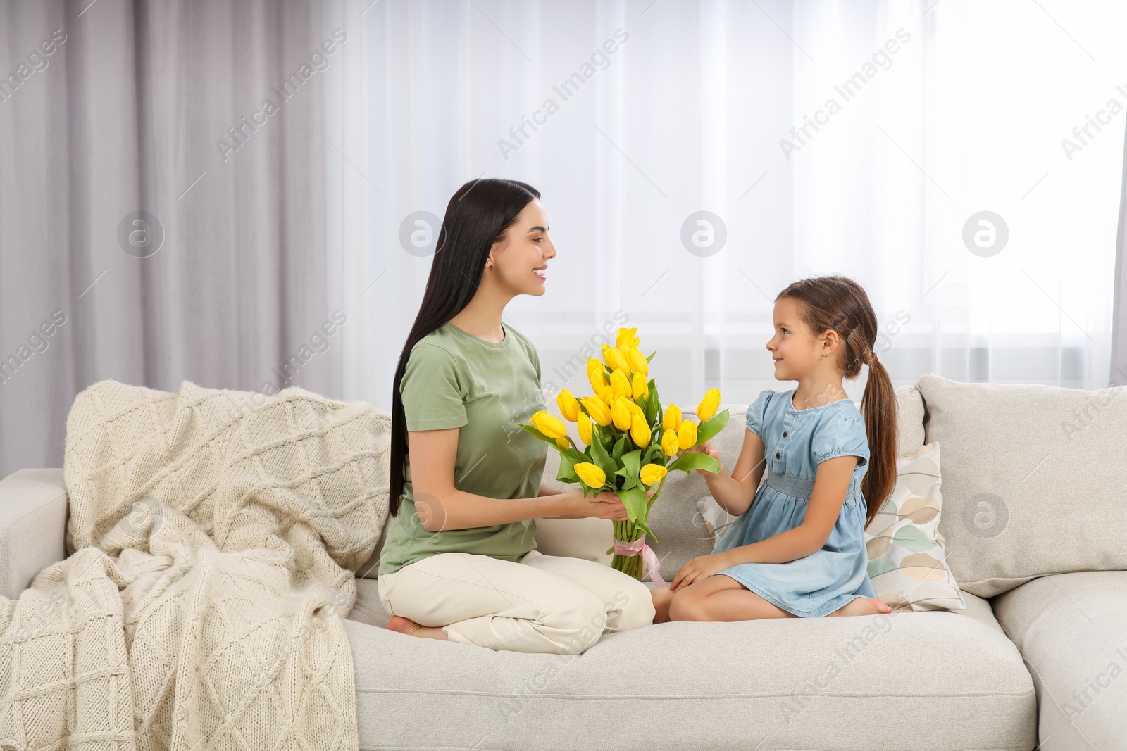 Photo of Happy woman and her cute daughter with bouquet of yellow tulips on sofa at home. Mother's day celebration