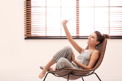 Photo of Young woman taking selfie near window with blinds at home. Space for text