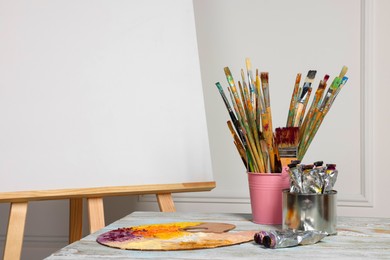 Photo of Easel with blank canvas and different art supplies on wooden table near white wall