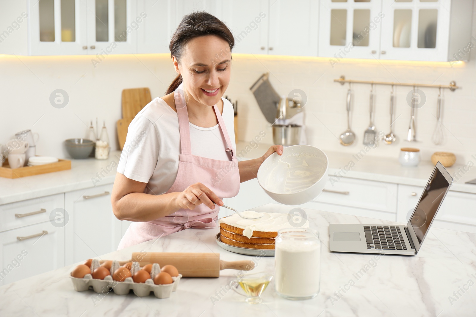 Photo of Woman making cake while watching online cooking course via laptop in kitchen