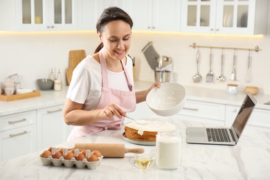 Photo of Woman making cake while watching online cooking course via laptop in kitchen