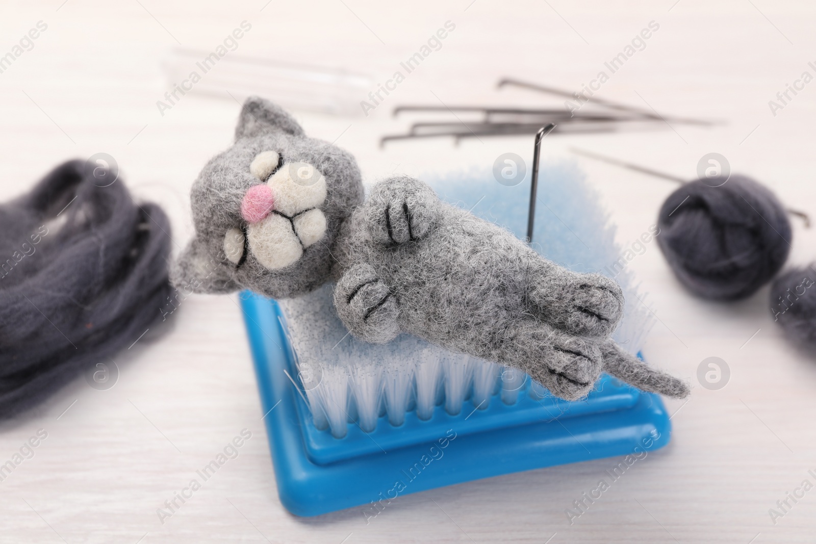 Photo of Felted cat, wool, needles and brush on light wooden table, closeup