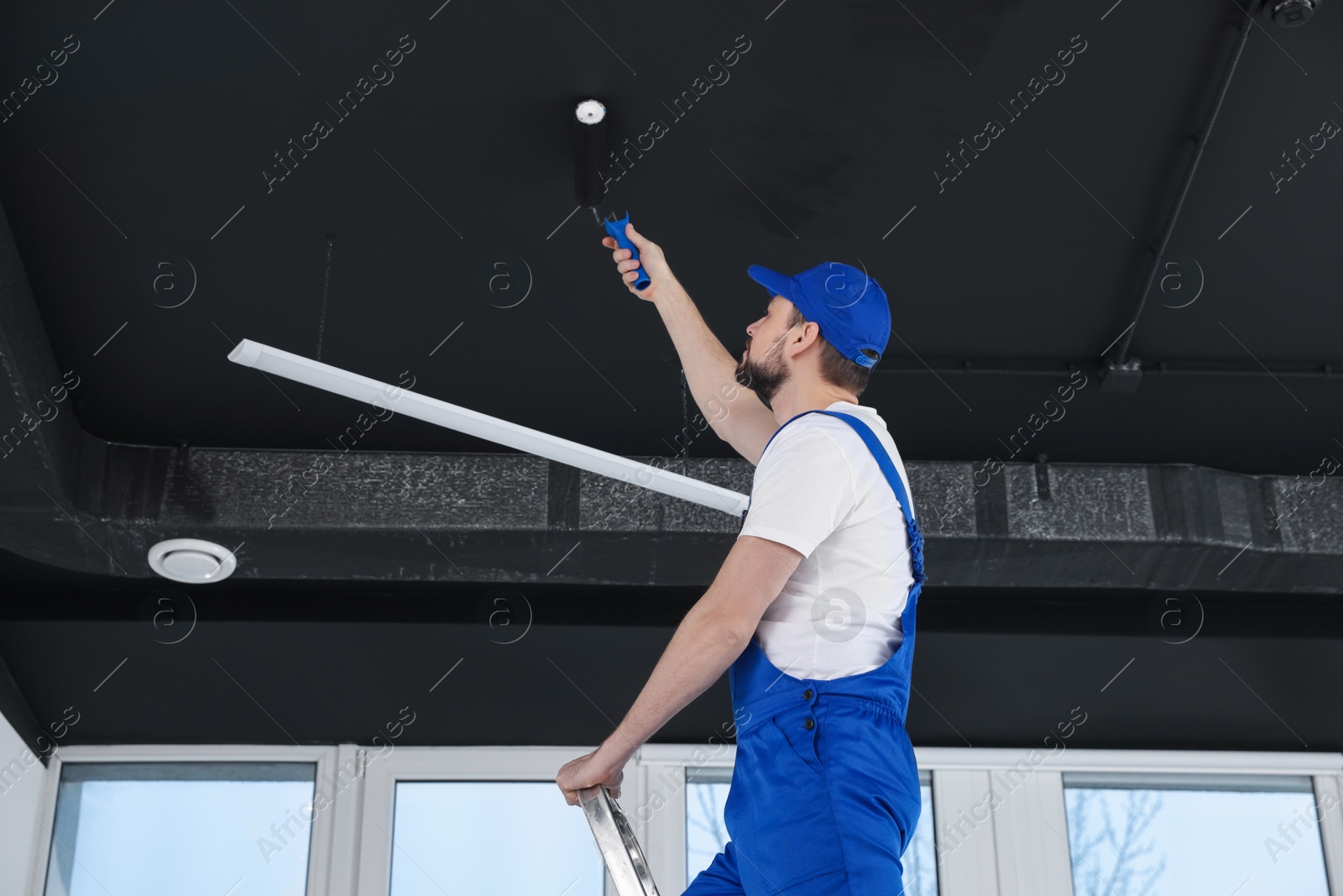 Photo of Worker in uniform painting ceiling with roller indoors