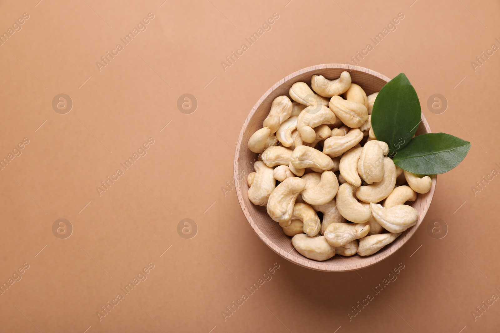 Photo of Tasty cashew nuts and green leaves in bowl on pale brown background, top view. Space for text