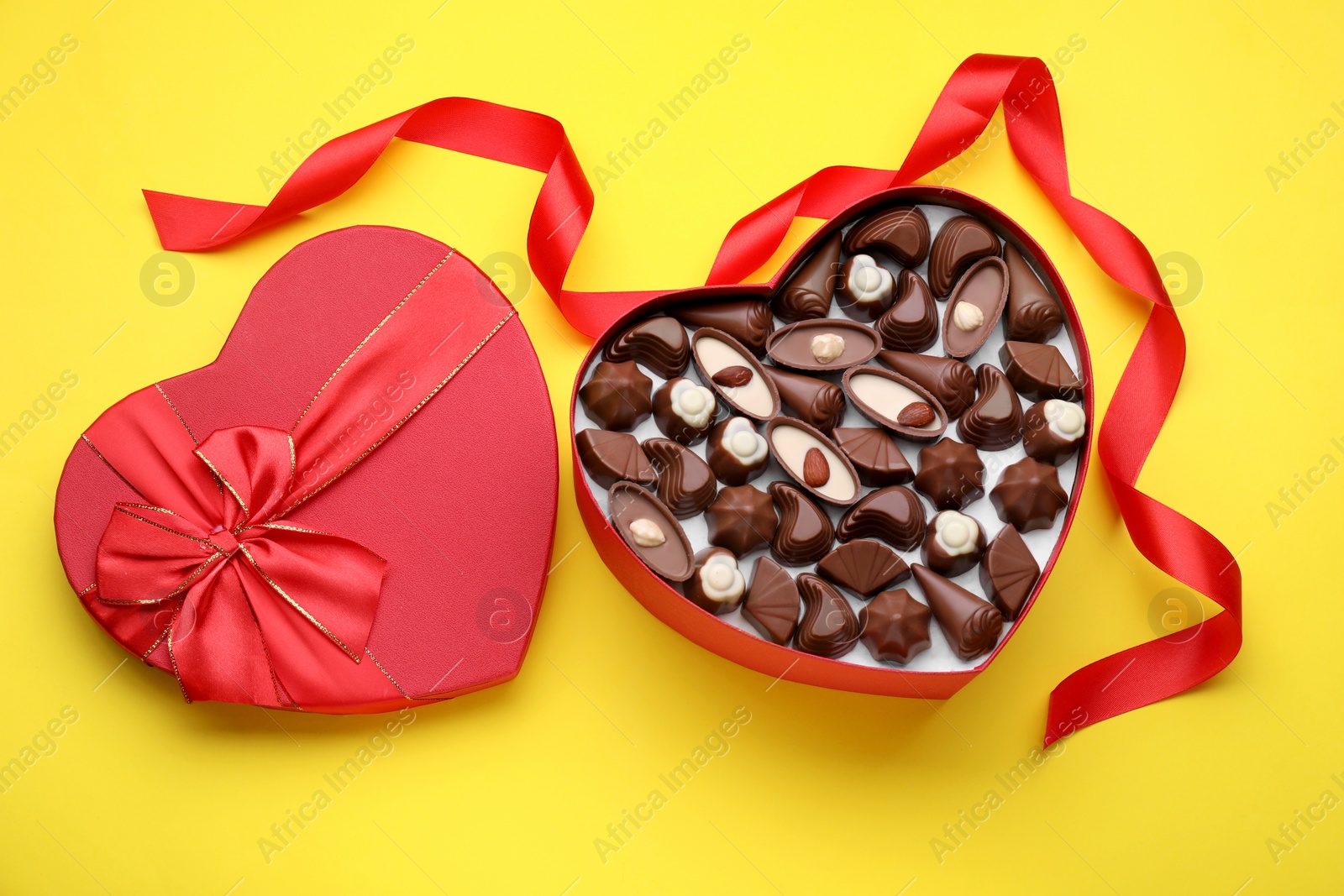 Photo of Heart shaped box with delicious chocolate candies and ribbon on yellow background, flat lay