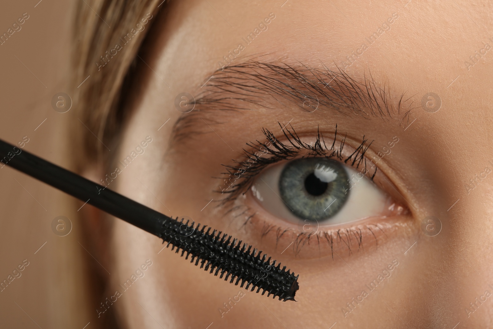 Photo of Woman applying mascara onto eyelashes against light brown background, closeup