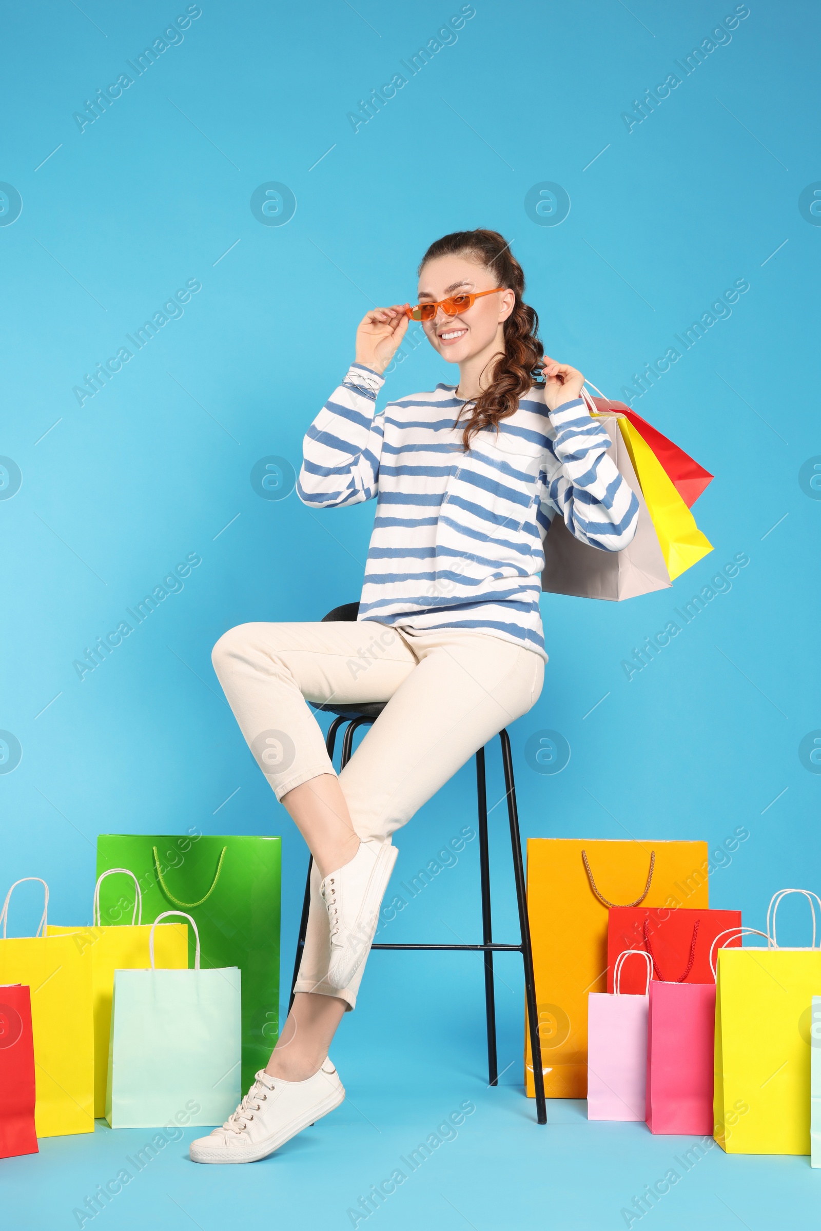 Photo of Happy woman in stylish sunglasses holding colorful shopping bags on stool against light blue background