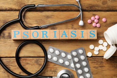 Photo of Word Psoriasis madecubes with letters, stethoscope and pills on wooden table, flat lay