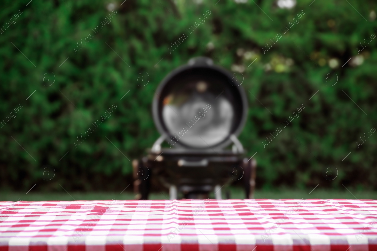 Photo of Picnic table with red checkered cloth and blurred barbecue grill outdoors