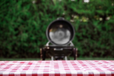 Photo of Picnic table with red checkered cloth and blurred barbecue grill outdoors