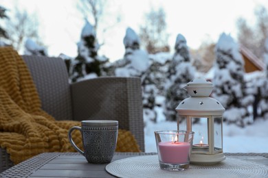 Burning candle, lantern and cup with hot drink on coffee table outdoors. Cosy winter