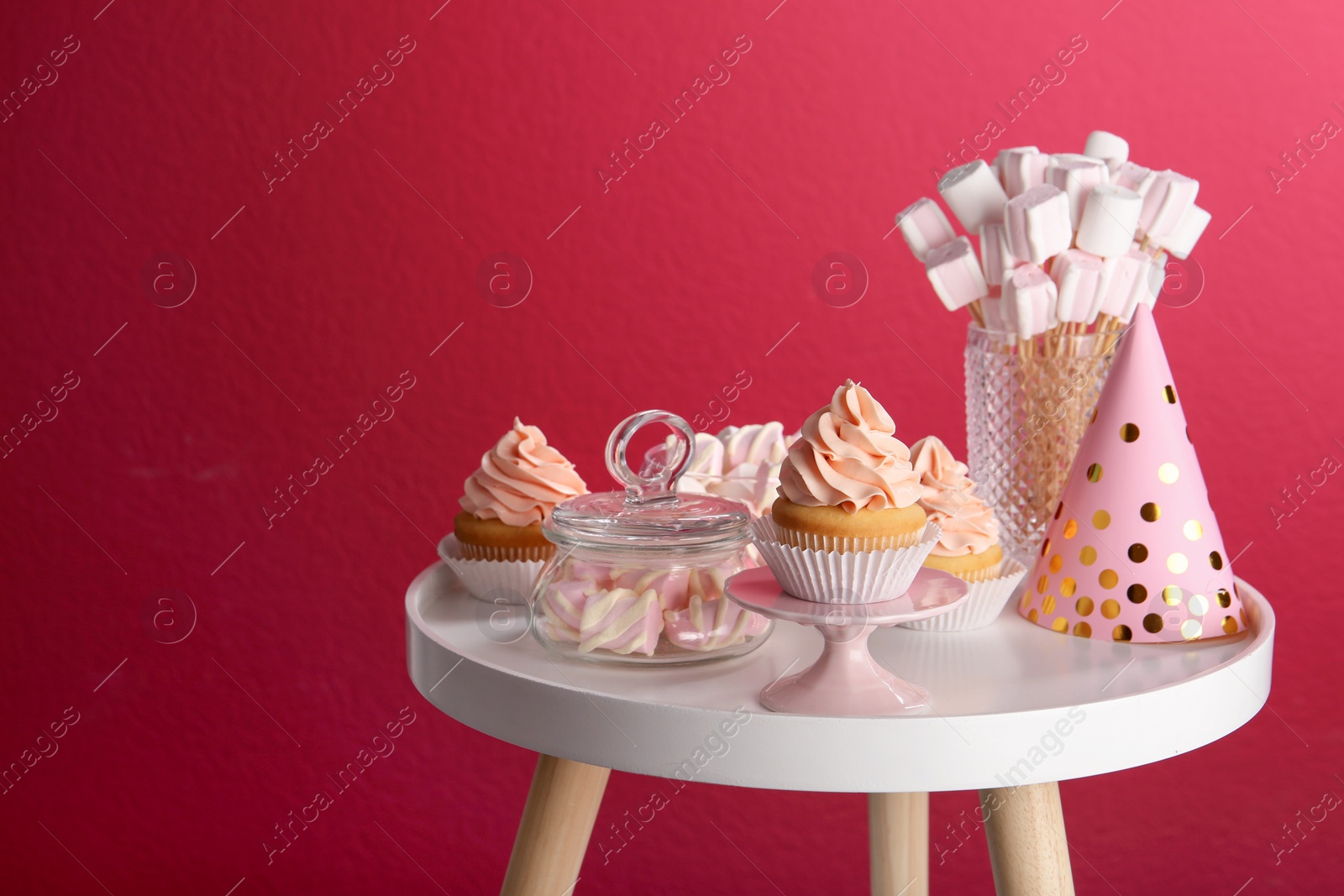 Photo of Table with party hat, cupcake and other sweets on burgundy background, space for text. Candy bar