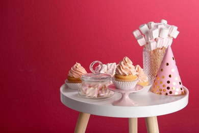 Table with party hat, cupcake and other sweets on burgundy background, space for text. Candy bar