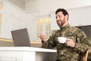 Photo of Happy soldier with cup of drink using video chat on laptop at white table in kitchen. Military service