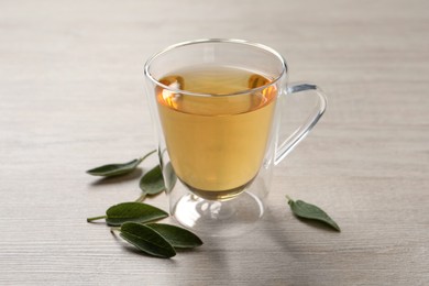 Photo of Glass cup of sage tea and green leaves on wooden table