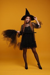 Photo of Young woman in scary witch costume with broom on orange background. Halloween celebration