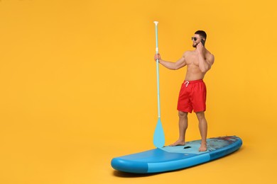 Happy man with paddle talking on smartphone on SUP board against orange background, space for text