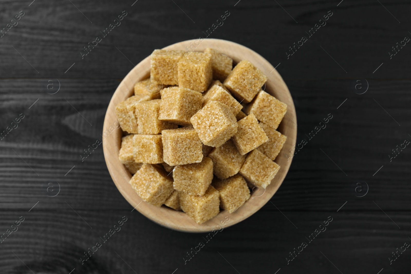 Photo of Brown sugar cubes in bowl on black wooden table, top view