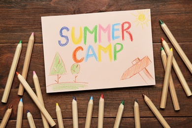 Card with text SUMMER CAMP, drawings and colorful pencils on wooden table, flat lay