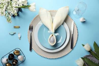 Photo of Festive table setting with painted eggs, plates and white tulips on light blue background, flat lay. Easter celebration