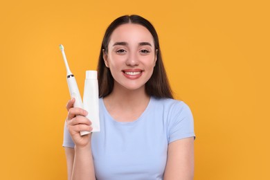 Photo of Happy young woman holding electric toothbrush and tube of toothpaste on yellow background