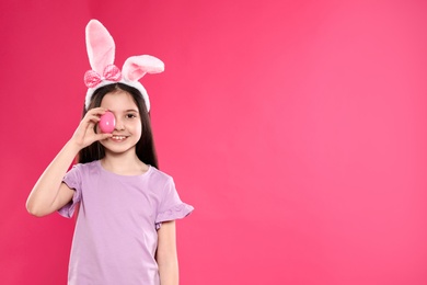 Little girl in bunny ears headband holding Easter egg near eye on color background, space for text