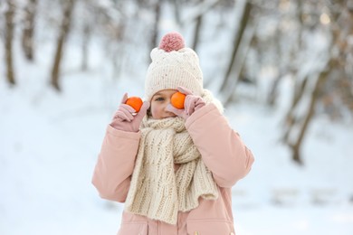 Cute little girl covering eye with tangerine in snowy park on winter day
