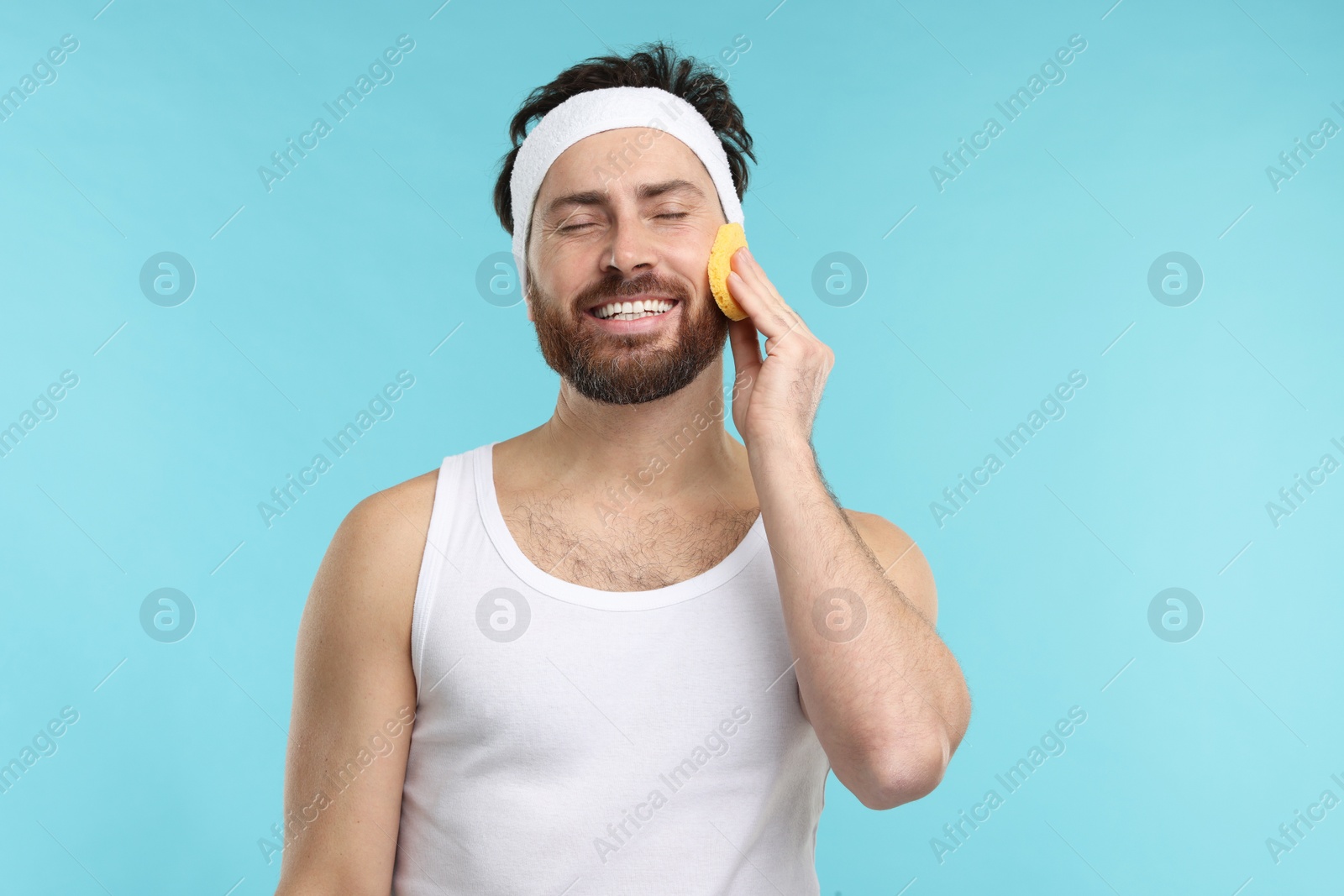 Photo of Man with headband washing his face using sponge on light blue background