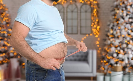 Overweight man measuring his waist in room with Christmas trees after holidays, closeup