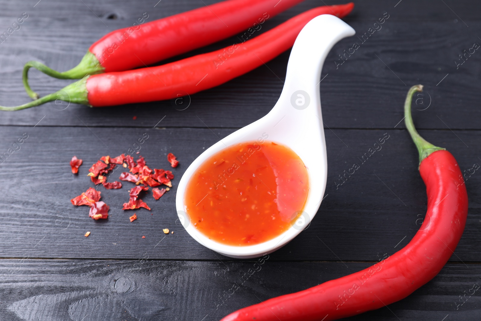 Photo of Spicy chili sauce in spoon and peppers on black wooden table