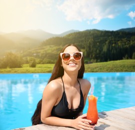 Image of Beautiful young woman with cocktail in outdoor swimming pool at luxury resort and beautiful view of mountains on sunny day