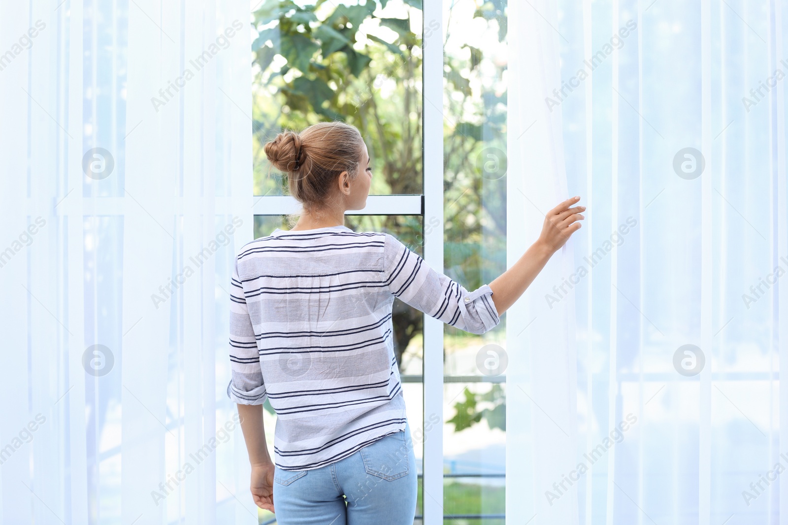 Photo of Young woman opening curtains and looking out of window at home