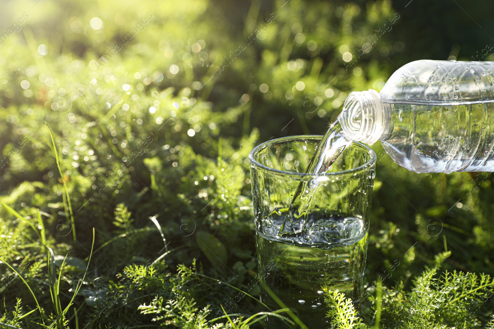 Photo of Pouring fresh water from bottle into glass on green grass outdoors. Space for text