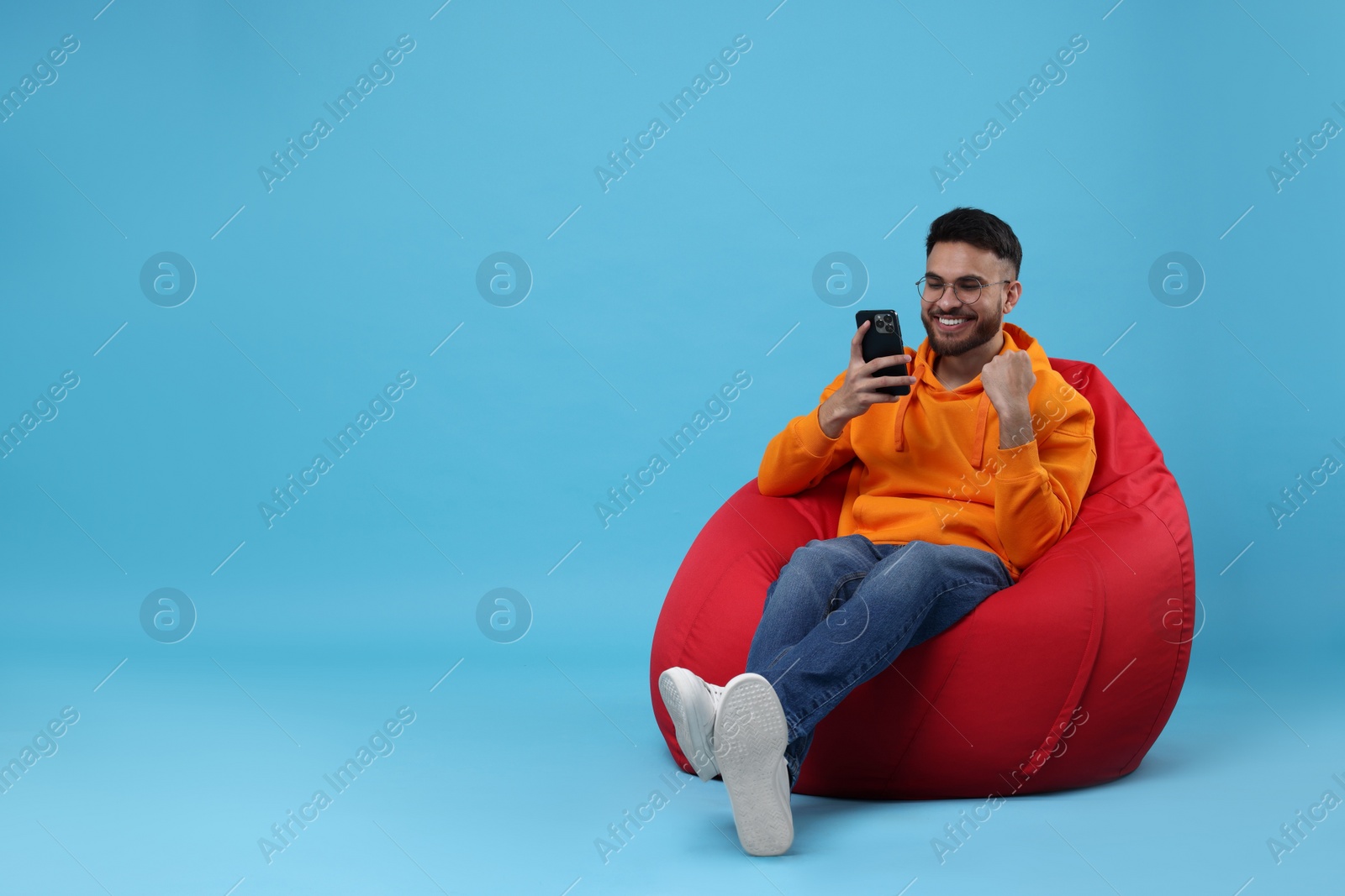 Photo of Happy young man using smartphone on bean bag chair against light blue background. Space for text