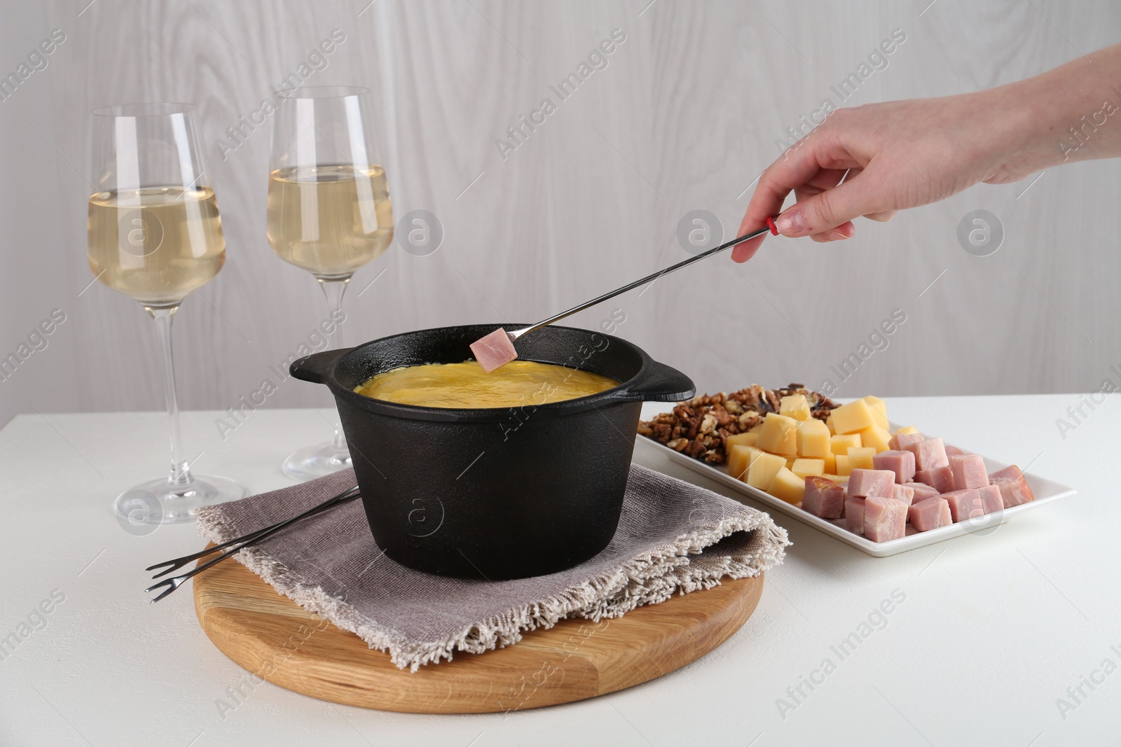 Photo of Dipping piece of ham into fondue pot with tasty melted cheese at white table, closeup