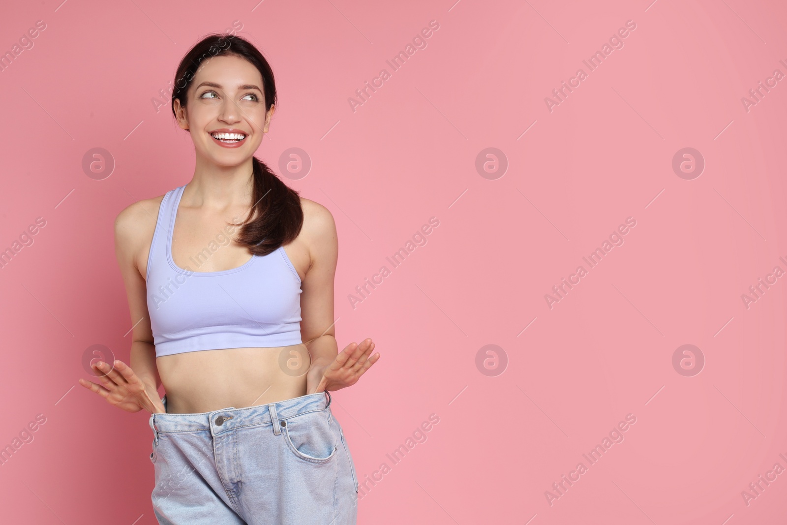 Photo of Happy young woman in big jeans showing her slim body on pink background, space for text