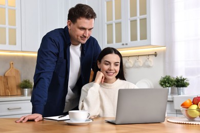 Photo of Happy couple using laptop together at wooden table in kitchen