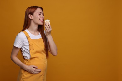 Pregnant young woman eating tasty cupcake on orange background, space for text