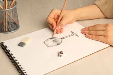 Woman drawing glass of wine with graphite pencil at light wooden table, closeup