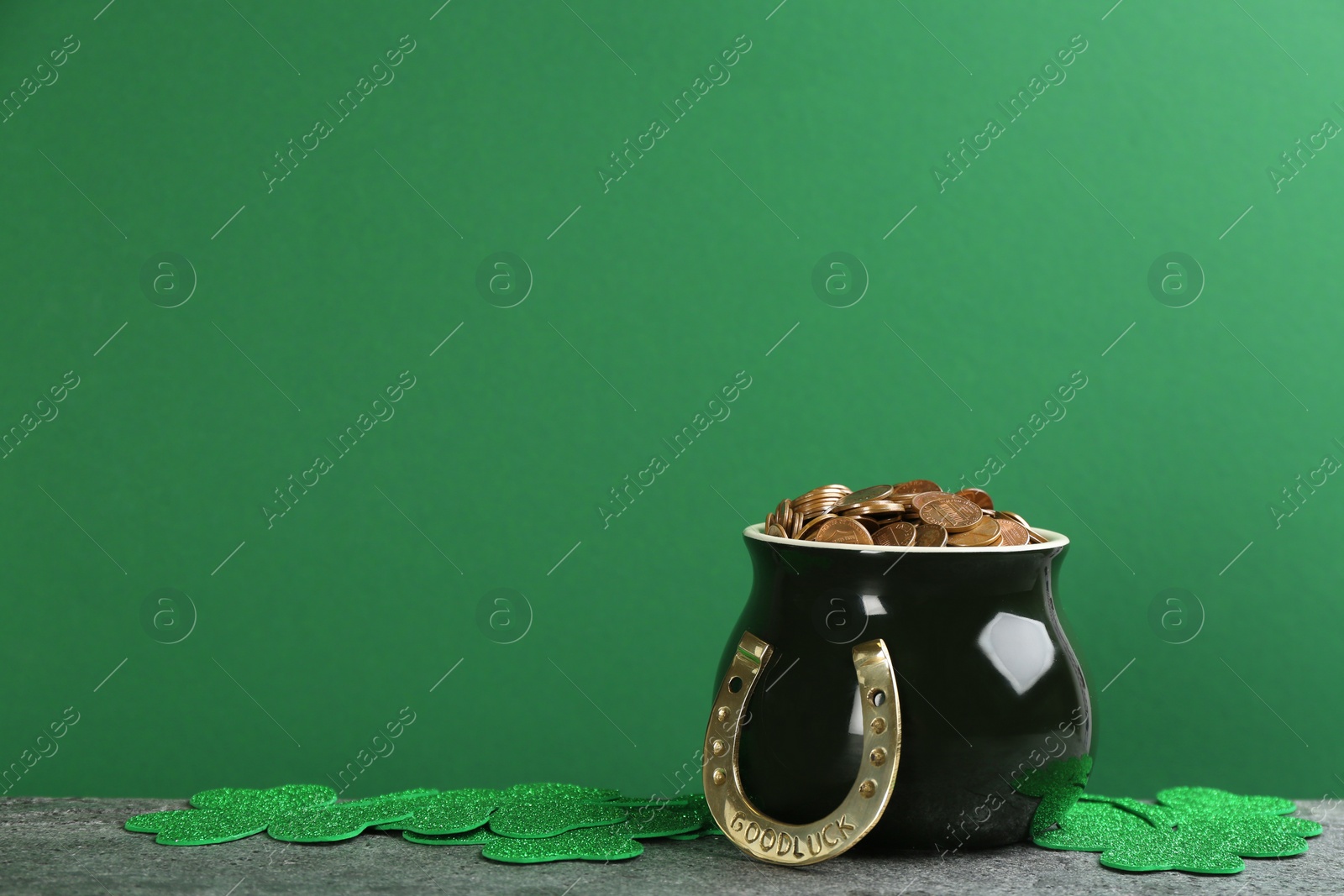 Photo of Pot of gold coins, horseshoe and clover leaves on grey stone table against green background, space for text. St. Patrick's Day celebration