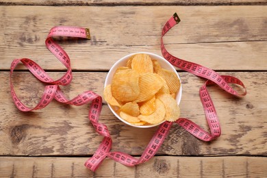 Bowl with potato chips and measuring tape on wooden table, flat lay. Weight loss concept