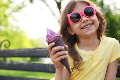 Cute little girl with delicious ice cream on bench in park, space for text