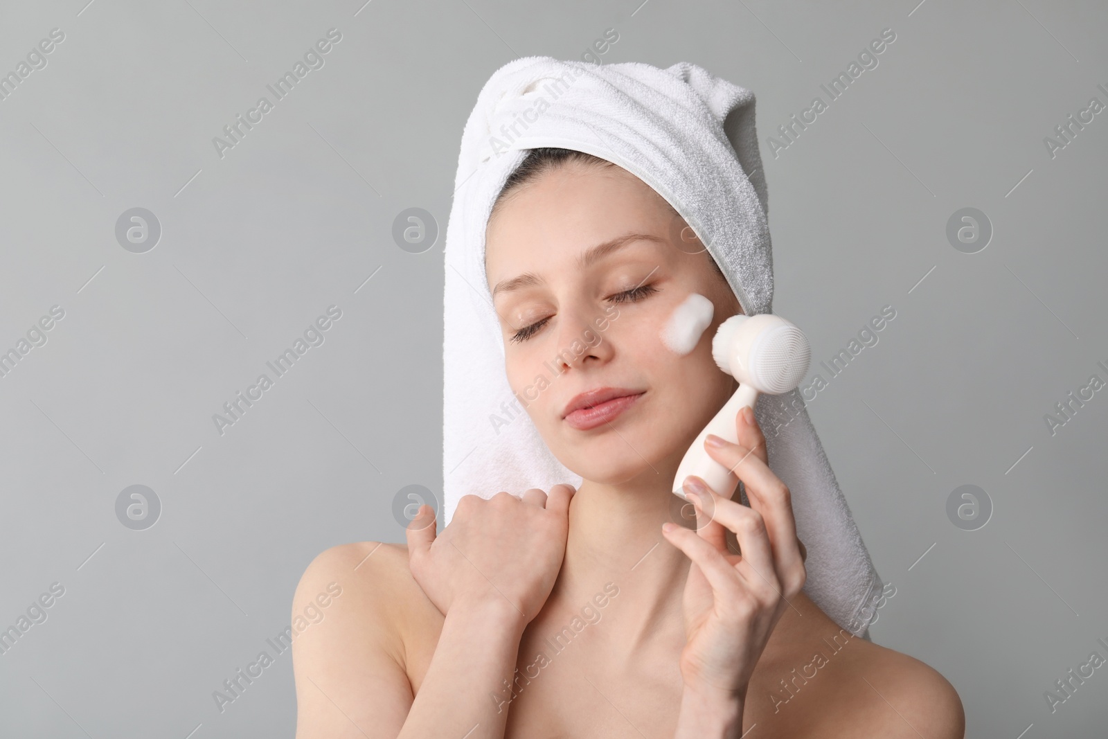 Photo of Young woman washing face with brush and cleansing foam on grey background