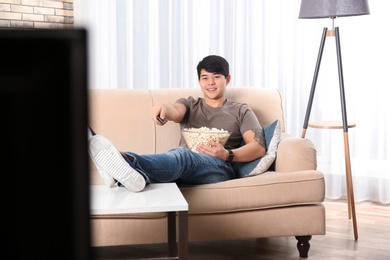 Young man with remote control and bowl of popcorn watching TV on sofa at home