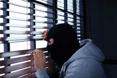 Masked man spying through window blinds indoors. Criminal offence