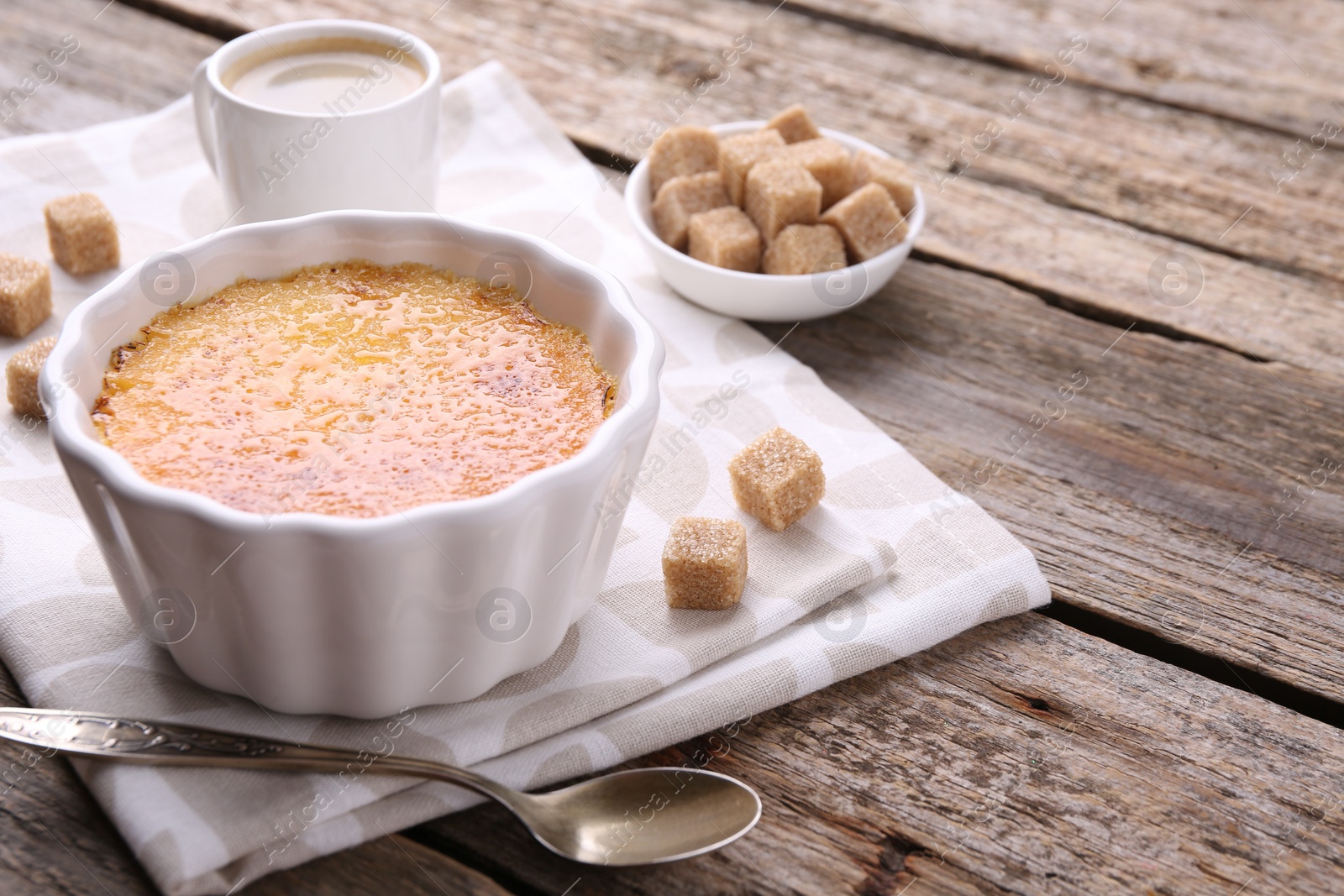 Photo of Delicious creme brulee in bowl, sugar cubes and spoon on wooden table, closeup