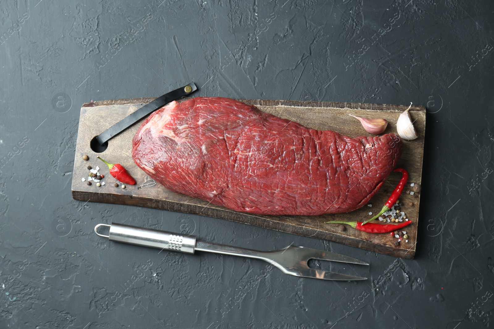 Photo of Piece of raw beef meat, garlic, chili, spices and fork on grey textured table, flat lay